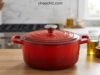Le Creuset Dutch Oven Review: Why Bakers Pay a Premium A vibrant Cerise red Le Creuset Signature enameled cast iron Dutch oven sitting on a wooden cutting board, with its tight-fitting lid resting beside it to reveal a deeply browned, crusty sourdough boule resting inside the pristine light-sand interior.