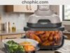 Ninja Crispi 4-in-1 Glass Air Fryer Review: The Death of the Plastic Basket A professional product photograph of the Ninja Crispi system on a modern kitchen counter. The 1500W PowerPod is shown mounted atop a clear, heavy-duty borosilicate glass container filled with crispy roasted vegetables. The image emphasizes the complete absence of plastic and PFAS-coated surfaces. A website watermark is positioned at the very top center of the frame.